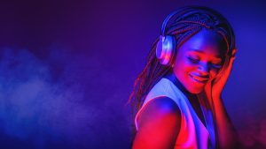 A person with braided hair smiles while wearing headphones in colorful lighting