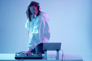 Person in a hoodie with headphones working on a DJ mixer and laptop in soft lighting